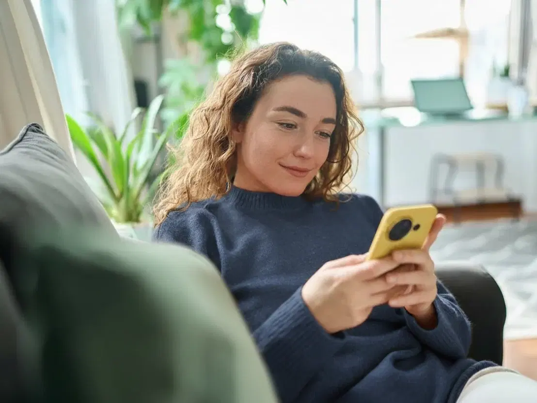 Lächelnde Frau, die auf ihrer Couch auf ihr Handy schaut.